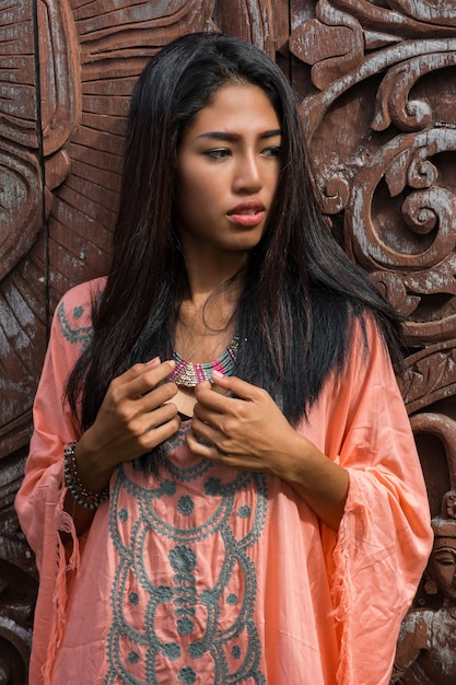 Bello modello asiatico in vestito rosa boho che posa sopra la parete ornamentale di legno.