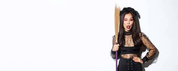 Free photo beautiful asian woman celebrating halloween in witch costume holding broom and showing tongue winkin