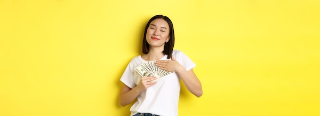 Beautiful asian woman love money hugging dollars and smiling pleased standing over yellow background