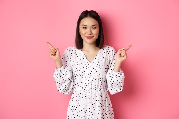 Beautiful asian woman making choice on shopping, pointing fingers sideways and showing variants, smiling at camera, standing over pink background