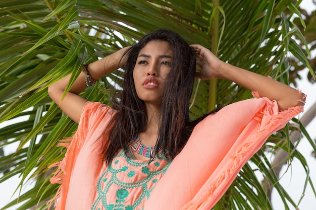 Beautiful asian woman posing on the tropical plants and leaves. Wearing trendy boho beach dress with embroidery and tassel. Jewelry , bracelet and necklace.