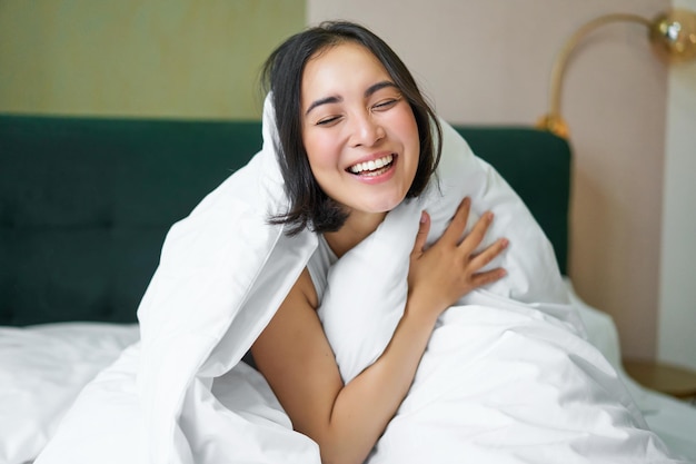 Foto gratuita bella donna asiatica che si siede sul letto coperto di piumino bianco che sorride godendo la mattina felice di fine settimana