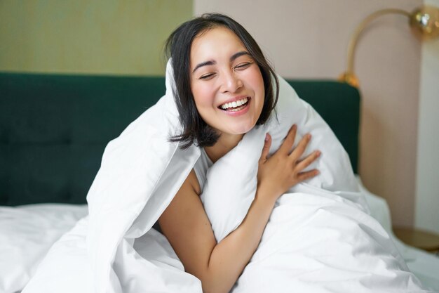 Beautiful asian woman sitting on bed covered with white duvet smiling enjoying happy weekend morning