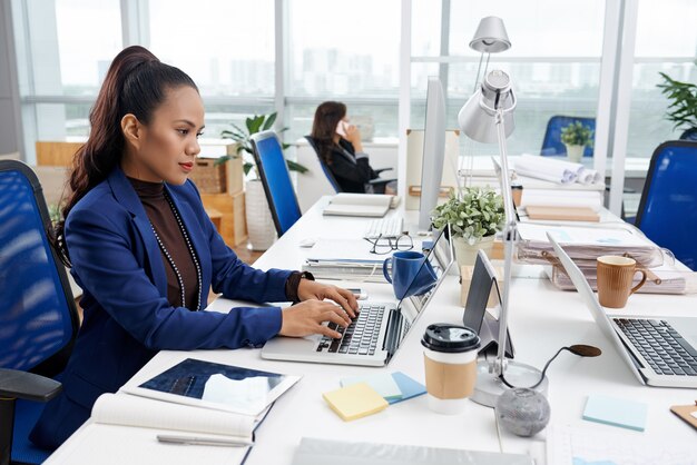 Beautiful Asian woman sitting at desk in busy office and working on laptop