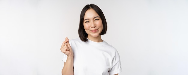 Beautiful asian woman smiling showing finger hearts gesture wearing tshirt standing against white ba