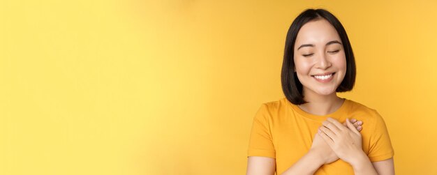 Beautiful asian woman smiling with tenderness and care holding hands on heart standing in tshirt ove