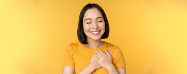 Beautiful asian woman smiling with tenderness and care holding hands on heart standing in tshirt ove