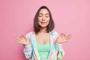 Бесплатное фото Красивая азиатская женщина стоит в позе дзен-нирвана с закрытыми глазами, практикует йогу, имеет позитивные мысли, дышит спокойствием и расслабляется, наслаждается мирной атмосферой, носит красочную рубашку, позирует в помещении