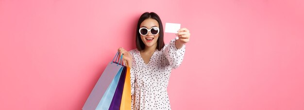 Beautiful asian woman in sunglasses going shopping holding bags and showing credit card standing ove