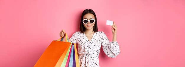Красивая азиатская женщина в солнечных очках ходит по магазинам, держа сумки и показывая кредитную карту, стоящую над