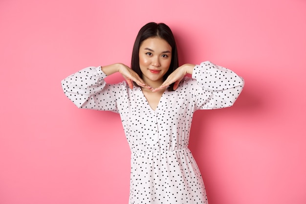 Beautiful asian woman in trendy dress making cute face, holding hands near jawline and gazing coquettish at camera, standing over pink background