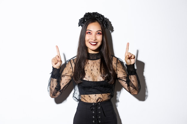Bella donna asiatica in costume da vedova non morta, indossa un abito di pizzo nero per halloween, punta il dito verso l'alto e sorride compiaciuta, guardando il tuo logo, in piedi su sfondo bianco.