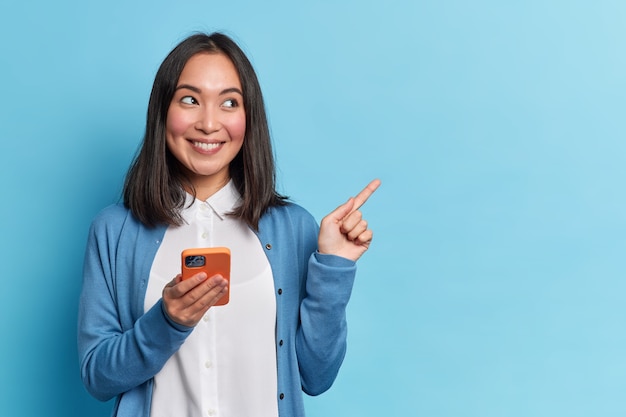 Beautiful Asian woman uses smartphone app sends messages in social media chat points away on copy space wears casual jumper