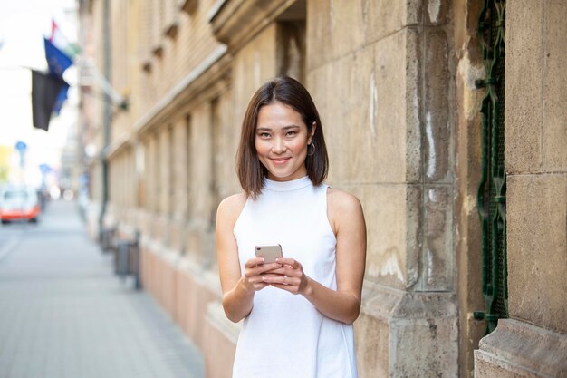 스마트폰으로 SMS 메시지를 보내기 위해 애플리케이션을 사용하는 아름다운 아시아 여성 거리에 스마트폰이 서 있는 행복한 젊은 아시아 여성