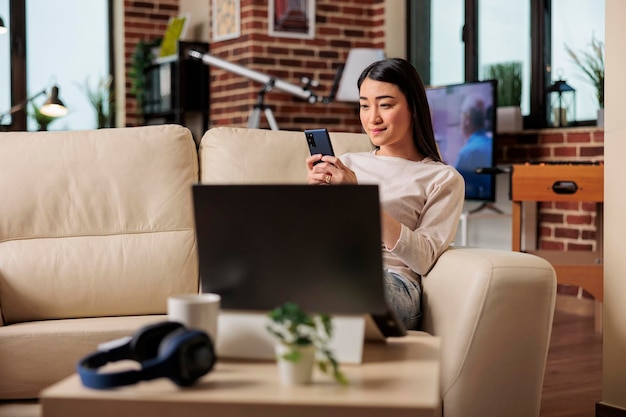 Bella donna asiatica che utilizza la tecnologia domestica del telefono, utente mobile online web. felice giovane donna seduta casualmente sul divano con lo smartphone in mano