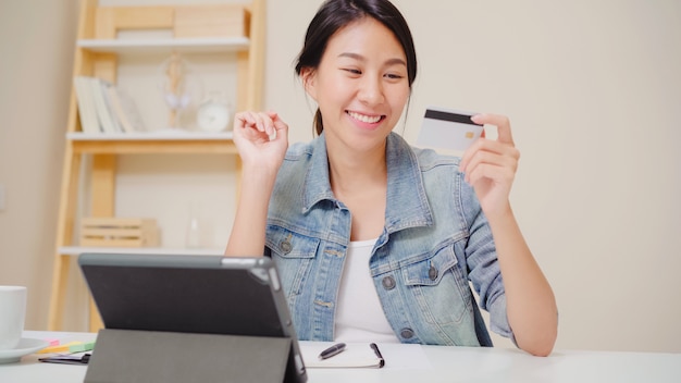 Beautiful Asian woman using tablet buying online shopping by credit card while wear casual sitting on desk in living room at home. 