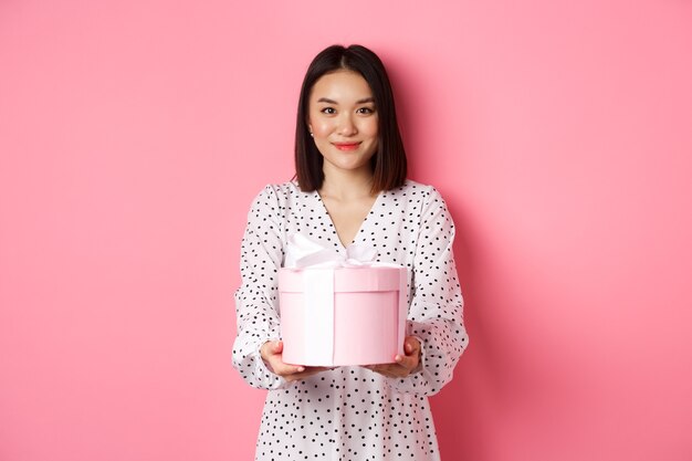 분홍색 배경에 서 있는 귀여운 상자에 선물을 주는 행복한 휴가를 바라는 아름다운 아시아 여성