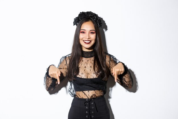 魔女の衣装とゴシック様式の化粧をした美しいアジアの女性、指を下に向け、ハロウィーンや死の日についてのプロモーションバナーを表示し、白い壁の上に立っています