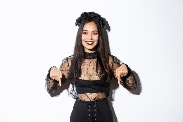 Beautiful asian woman in witch costume and gothic style makeup, pointing fingers down, showing promo banner about halloween or day of death, standing over white wall