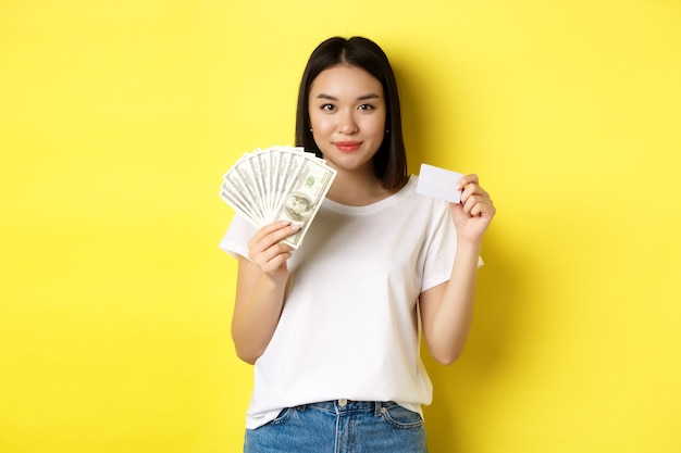 짧은 검은 머리를 가진 아름다운 아시아 여성, 흰색 티셔츠를 입고, 달러와 플라스틱 신용 카드로 돈을 보여주고, 노란색 배경 위에 서 있습니다.
