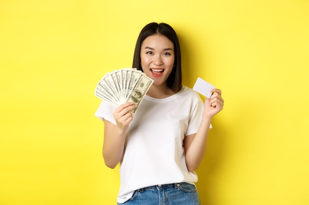 短い​黒髪​、​白い​tシャツ​を​着て​、​ドル​と​プラスチック​の​クレジットカード​で​お金​を​示し​、​黄色​の​背景​の​上に​立っている​美しい​アジア​の​女性