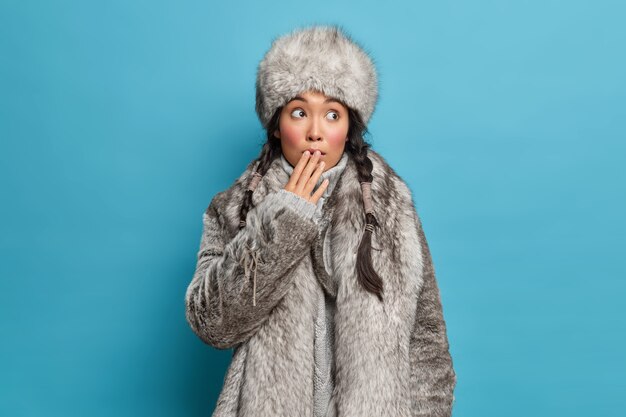 Бесплатное фото Красивая азиатская женщина с двумя косичками прикрывает рот и чувствует себя потрясенной, носит теплую шубу из натурального меха и платья шляпы для холодной погоды, живет на севере, изолированном от синей стены