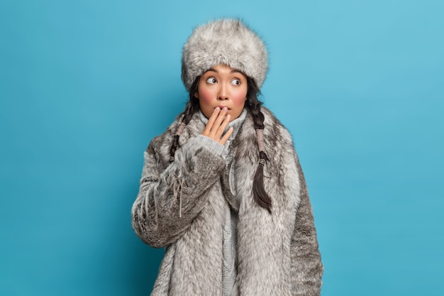 Beautiful Asian woman with two pigtails covers mouth and feels shocked wears warm natural fur coat and hat dresses for cold weather lives at north isolated over blue wall