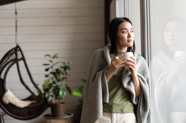 담요에 싸인 아름다운 아시아 여성이 창가에 기대어 뜨거운 차를 마시고 밖을 바라보고 있다