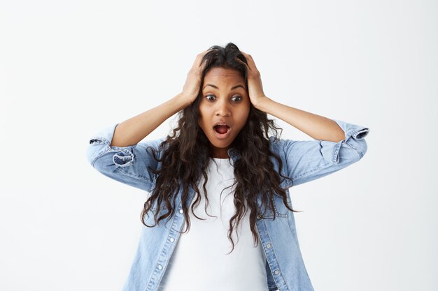 Beautiful astonished African-american woman wearing loose wavy black hair holding hands on her head having wide opened eyes and mouth being shocked to hear horror news.