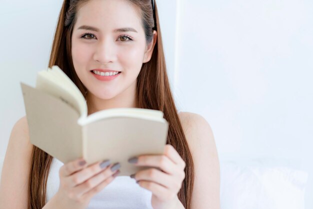 아름다운 매력적인 아시아 여성은 침대에서 책을 읽는 것을 즐깁니다. 아시아 긴 머리 여성의 초상화는 주말 활동 흰색 침실을 즐깁니다.