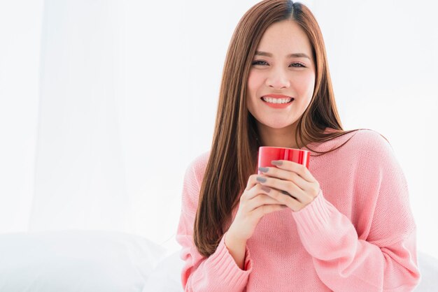 Красивая привлекательная азиатская женщина в розовом свитере с горячим напитком на белой кровати, утренняя свежесть, здоровый образ жизни