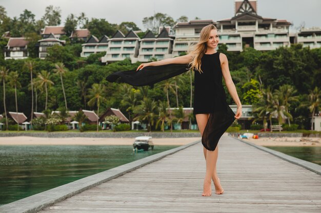 Beautiful attractive woman dressed in black dress posing on pier in luxury resort hotel, summer vacation, tropical beach