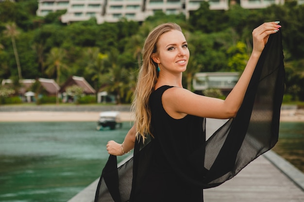 Beautiful attractive woman dressed in black dress posing on pier in luxury resort hotel, summer vacation, tropical beach