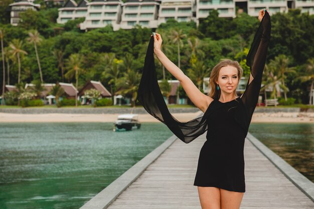 Beautiful attractive woman dressed in black dress posing on pier in luxury resort hotel, summer vacation, tropical beach