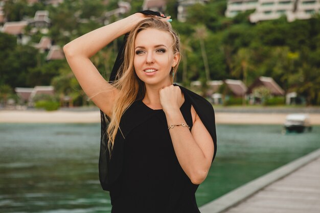 Beautiful attractive woman dressed in black dress posing on pier in luxury resort hotel, summer vacation, tropical beach