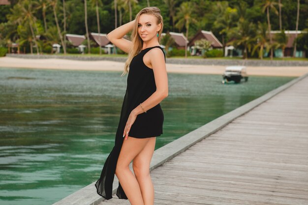 Beautiful attractive woman dressed in black dress posing on pier in luxury resort hotel, summer vacation, tropical beach