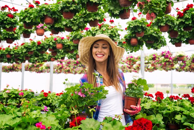 Beautiful attractive woman florist with hat walking through greenhouse holding potted flowers and checking plants