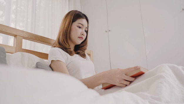 Beautiful attractive young Asian woman reading a book while lying on the bed 