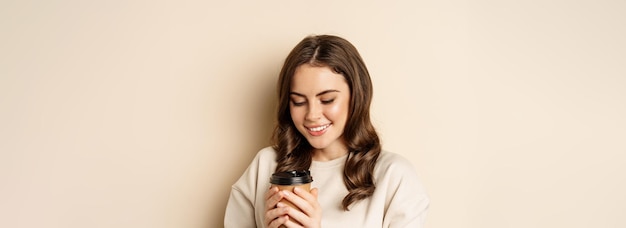 Бесплатное фото Красивая аутентичная женщина улыбается, держа теплую чашку кофе и выглядя счастливой, стоя над бежевым б