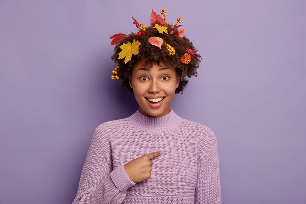 아름다운 가을 소녀는 계절 축제에 참여하기 위해 픽업되어 기뻐하며 따뜻한 니트 스웨터, 화려한 잎, 열매 및 꽃을 머리카락에 착용하고 보라색 배경에 고립 된 자신을 가리 킵니다.