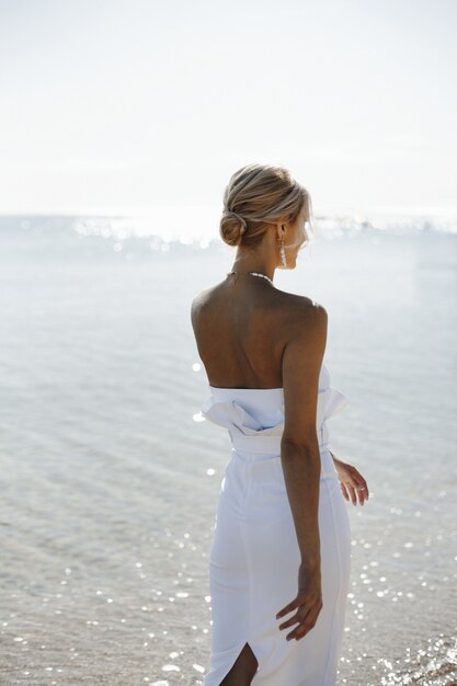 Beautiful back view of a young blonde woman in white dress is walking near the sea on the sunny day