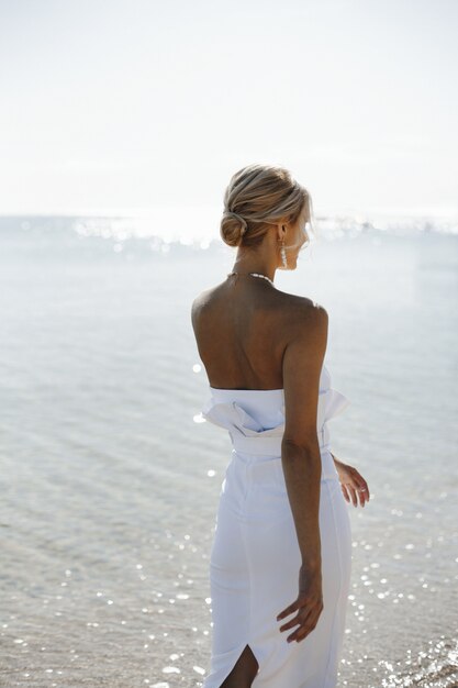 白いドレスの若いブロンドの女性の美しい背面図は、晴れた日に海の近くを歩いています。
