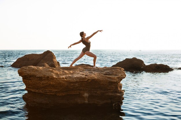 Красивая балерина танцует, позирует на скале на пляже, вид на море.