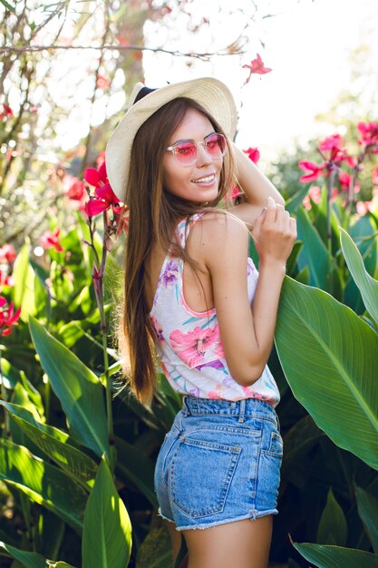 Красивая пляжная девушка, стоящая в парке в соломенной шляпе, розовых очках, джинсовых шортах и яркой футболке. Девушка очень стройная и привлекательная. Она выглядит игривой. Она стоит среди тропических цветов