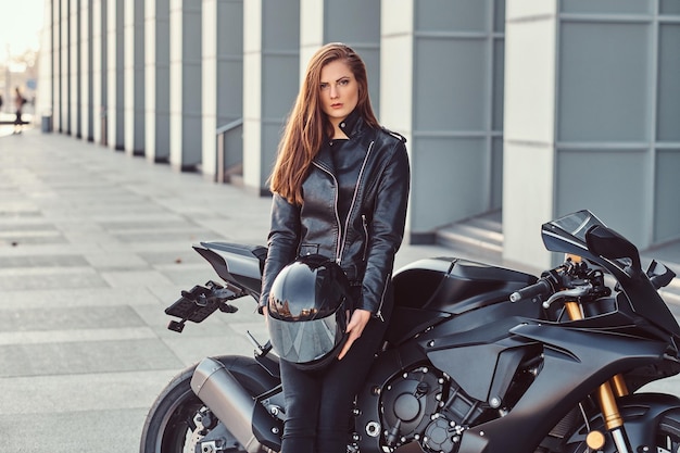 A beautiful biker girl wearing black leather jacket holding helmet while leaning on her superbike outside a building.