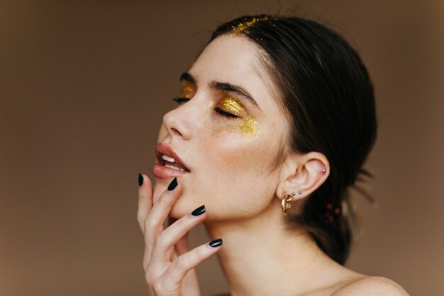 Beautiful black-haired girl posing with eyes closed. Close-up shot of blissful caucasian lady isolated on brown wall.