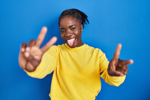파란색 배경 위에 서 있는 아름다운 흑인 여성은 승리의 표시를 하고 있는 두 손의 손가락을 내보이며 혀를 내밀고 웃고 있습니다. 두 번째.