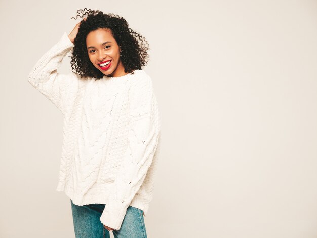 Beautiful black woman with afro curls hairstyle and red lips.Smiling model in  trendy jeans clothes and winter sweater. 