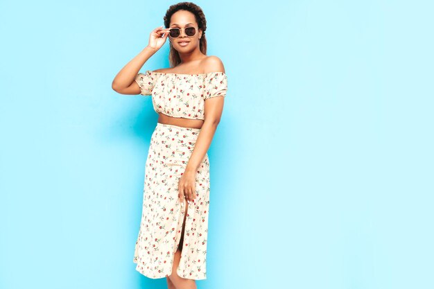 Beautiful black woman with afro curls hairstyle Smiling model dressed in summer dress Sexy carefree female posing near blue wall in studio Tanned and cheerful In sunglasses