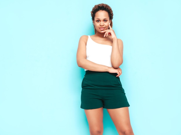 Free photo beautiful black woman with afro curls hairstyle smiling model dressed in summer shorts sexy carefree female posing near blue wall in studio tanned and cheerful